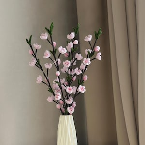 Op de afbeelding: Een crèmekleurige vaas met een verticale geribbelde textuur bevat een boeket kunstmatige kersenbloesemtakken. De takken zijn donkerbruin met kleine groene blaadjes en talloze lichtroze bloemen met elk vijf bloemblaadjes, in een hoge, rechtopstaande opstelling.