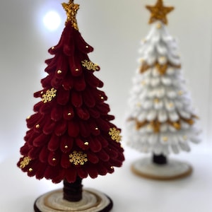 May include: Two decorative Christmas trees. One is burgundy with gold snowflake and bead accents, topped with a gold star. The other is white with gold accents and a gold star. Both trees are on round bases.