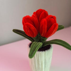 Op de afbeelding: Een decoratief kunstbloemstuk met levendige rode tulpen en groene bladeren, gepresenteerd in een witte pot met textuur. Een charmant accent voor de interieurdecoratie.