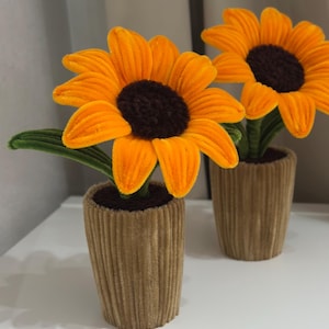 May include: Two plush sunflowers with bright orange petals and dark brown centres, each planted in a textured, ribbed tan pot. The flowers feature soft, velvet-like fabric petals and green stems with leaves, standing upright as decorative items.