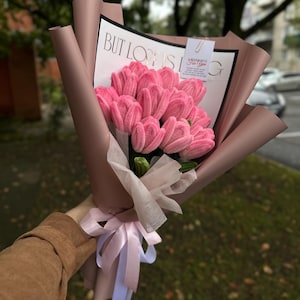 May include: A bouquet of pink crocheted tulips wrapped in mauve paper with a white ribbon. The bouquet includes a white card with the text BUT LOVE IS and a small tag that reads A LITTLE HUG FOR YOU. The flowers have a textured yarn appearance.