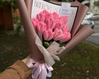 May include: A bouquet of pink crocheted tulips wrapped in mauve paper with a white ribbon. The bouquet includes a white card with the text BUT LOVE IS and a small tag that reads A LITTLE HUG FOR YOU. The flowers have a textured yarn appearance.