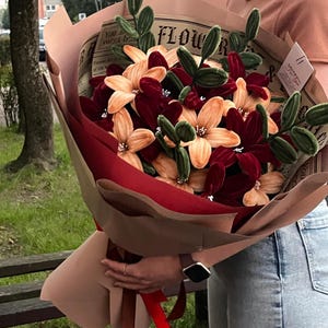 Puede incluir: Un ramo de flores artificiales envuelto en papel marrón y cinta roja. El ramo presenta flores de color burdeos y melocotón, con detalles verdes. El papel de envolver tiene el texto "FLOWER".