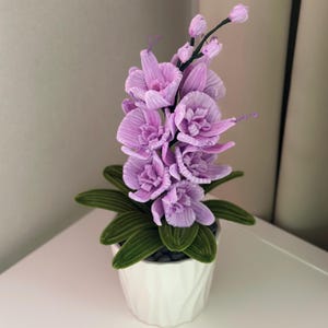 Pode incluir: Uma orquídea artificial em vaso com flores roxas claras e folhas verdes. A orquídea está em um vaso de cerâmica branco texturizado. O arranjo está em uma superfície branca.