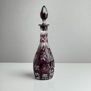 May include: A deep purple cut glass decanter featuring a clear glass stopper. The body of the decanter is decorated with intricate etched patterns of grape clusters and leaves, with a faceted neck and a starburst design on the base.