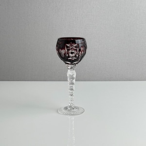 May include: A deep red cut-to-clear crystal wine glass featuring intricate starburst and floral patterns on the bowl. The stem is clear glass with a textured, faceted design, leading down to a circular, ribbed base with a clear glass finish.