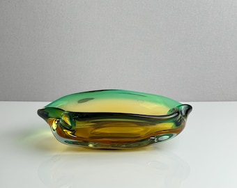 May include: A shallow, oval-shaped glass bowl featuring a smooth, translucent gradient transition from a vibrant emerald green at the upper rim to a warm, deep amber yellow at the base, with a polished, reflective surface and curved edges.