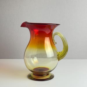 May include: A vintage glass pitcher with a vibrant color gradient. The top is a deep red, transitioning to orange and then yellow towards the base. It has a curved handle and a spout for pouring. The pitcher sits on a round base.