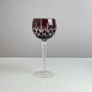 May include: A deep purple wine glass featuring a clear, textured stem and a round base with a radial pattern. The bowl of the glass is adorned with a diamond-cut geometric design that catches the light, contrasting with the clear glass stem.