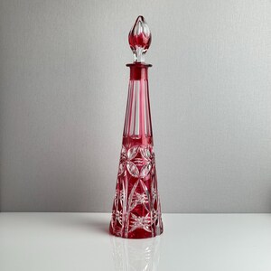 May include: A tall, conical glass decanter featuring a vibrant ruby red color with clear cut-glass accents. The body displays intricate geometric patterns, including starburst and circular motifs, topped with a matching pointed glass stopper.