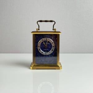 May include: A rectangular gold-tone carriage clock featuring a deep blue speckled face with a circular silver dial. The clock has black Roman numerals, gold hands, and a top carrying handle. The text "Quartz" is printed on the face.