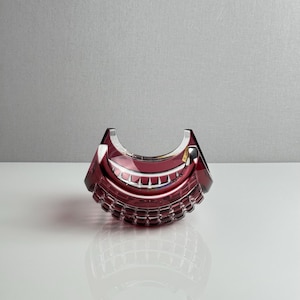 May include: A deep red, crescent-shaped crystal bowl featuring a faceted, geometric pattern along its curved exterior. The glass displays a rich, translucent burgundy hue with sharp, polished edges and a smooth, concave top opening.
