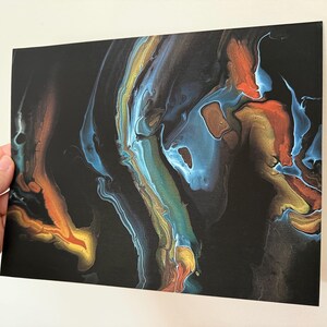 May include: A rectangular art print featuring an abstract acrylic pour design on a black background. The composition displays fluid, marbled streaks of metallic gold, vibrant orange, and cool blue tones that swirl across the dark surface.