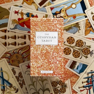 May include: A deck of "The Othrysian Tarot" cards in a box with a speckled orange and white design. The title is in a white rectangle. The deck is surrounded by other tarot cards with various illustrations.