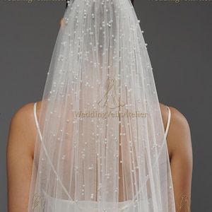 May include: A sheer white bridal veil, adorned with scattered pearl beads. The veil is draped over a person's head and shoulders, with the text "WeddingVeilsAtelier" visible in several places. The veil is a delicate accessory for weddings.
