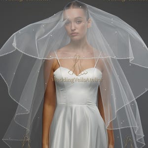 May include: A white bridal veil with a pearl beaded edge. The veil is draped over a white satin wedding dress. The text "WeddingVeilsAtelier" is visible in gold lettering on the image.