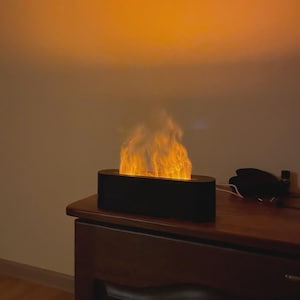 Peut inclure: Une cheminée électrique noire de forme ovale avec un effet de flamme simulé. Les flammes émettent une lueur orange chaude, créant une ambiance chaleureuse. La cheminée est posée sur une surface en bois sombre.