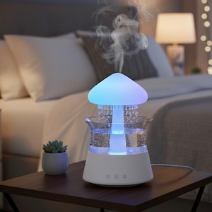 Peut inclure: Diffuseur d'huiles essentielles en forme de champignon blanc et transparent, avec un sommet lumineux bleu et violet. Le diffuseur est posé sur une table en bois brun foncé, à côté d'une petite plante succulente en pot. Le diffuseur émet une fine brume.