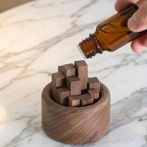 Peut inclure: Diffuseur d'huiles essentielles en bois de couleur marron foncé. Il se compose d'une base cylindrique et d'un assemblage de petits blocs de bois. Une bouteille en verre brun est tenue au-dessus, suggérant l'ajout d'huile.