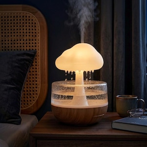 Può includere: Un diffusore di oli essenziali a forma di fungo con una parte superiore a forma di nuvola, che emette vapore. Il diffusore ha un serbatoio d'acqua in vetro trasparente e una base in legno. È su un comodino di legno.