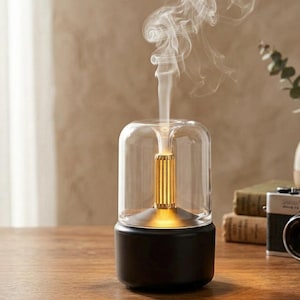 Peut inclure: Un diffuseur d'huiles essentielles noir et transparent avec un intérieur doré. Le diffuseur émet un flux de vapeur blanche. La base est noire et le dessus est en verre transparent. Le diffuseur est posé sur une surface en bois.