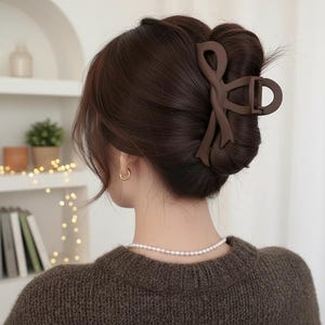 Könnte beinhalten: Eine braune Haarspange hält eine Hochsteckfrisur zusammen. Die Spange hat ein schleifenähnliches Design und ein mattes Finish. Die Person trägt einen braunen Pullover und eine Perlenkette. Goldene Creolen sind sichtbar.