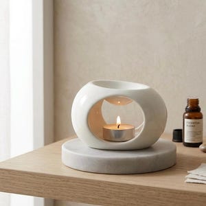 May include: A white ceramic oil burner with an oval shape and a central opening containing a lit tea light candle. The burner features a small basin at the top for oil. Beside it sits a small amber glass bottle labeled Essential Blend.