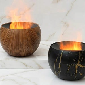 Peut inclure: Deux lampes décoratives à effet de flamme. L'une est en imitation bois, l'autre noire avec des veines dorées. Les deux ont une forme ronde et émettent une lueur orange, simulant des flammes. Elles sont posées sur une surface en marbre blanc.