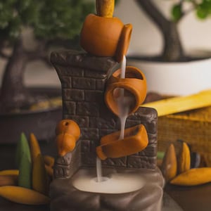 Puede incluir: Fuente de cascada de incienso de cerámica marrón con diseño de ladrillo. La fuente presenta múltiples niveles de jarrones vertedores, con vapor similar al humo que se eleva desde la base. Conos de incienso visibles.