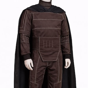 Puede incluir: Un disfraz marr&oacute;n con costuras blancas, con cuello alto, mangas largas y pantalones a juego. Una capa negra est&aacute; drapeada sobre los hombros. El disfraz tiene un dise&ntilde;o estructurado con cintur&oacute;n y detalles acolchados.
