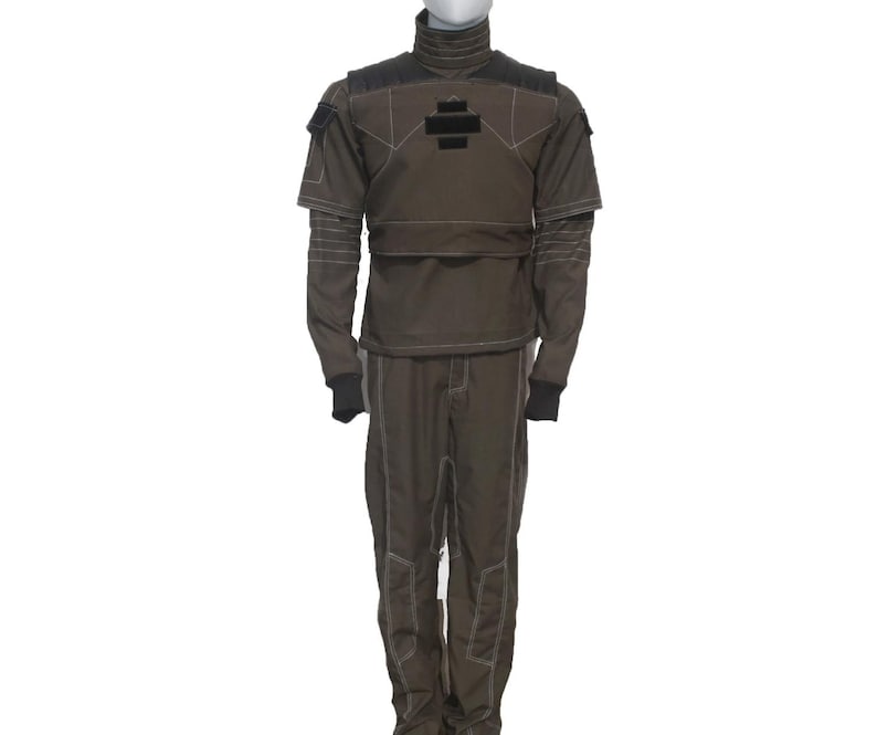 May include: A two-piece olive green jumpsuit with black accents and white stitching. The top has a high collar and a black patch with a cross design. The sleeves are long, and the pants are straight-legged.