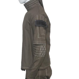 May include: Olive green jumpsuit with white stitching details. Features a high collar, black patch on the shoulder, and geometric patterns on the sleeves. The jumpsuit has long sleeves and pants with black cuffs.