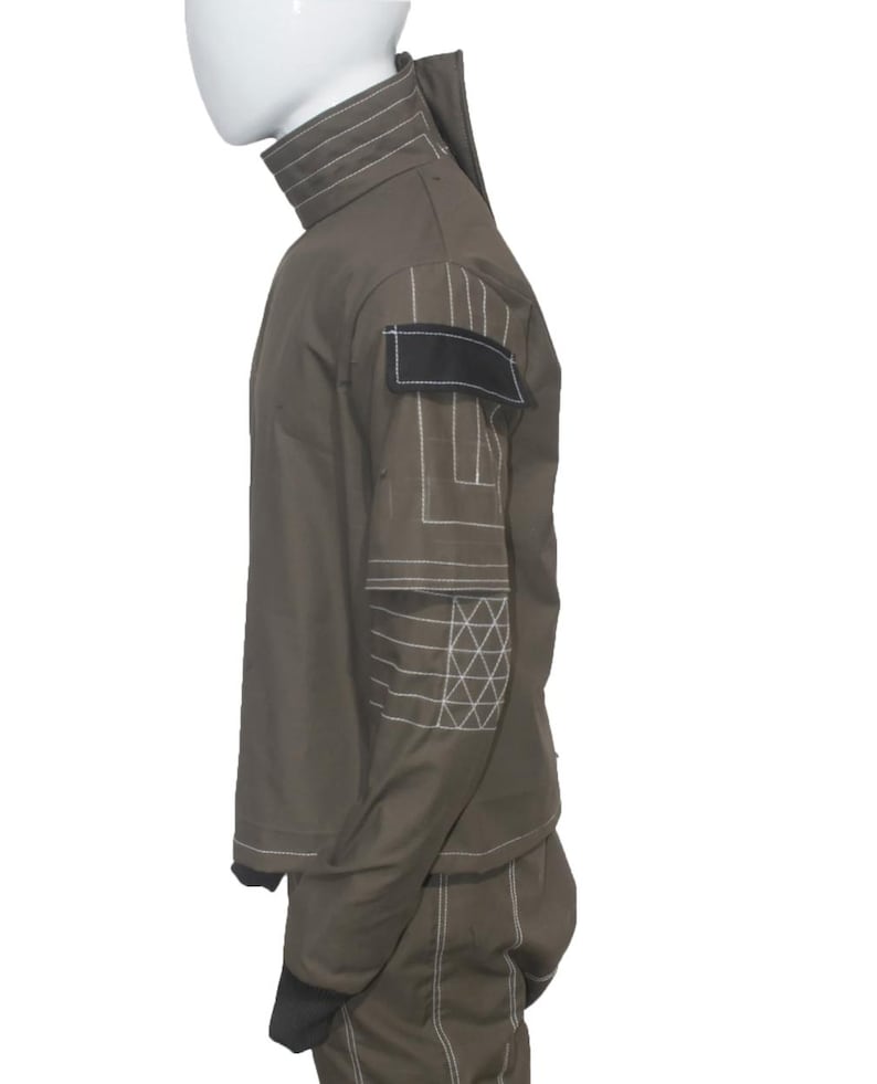 May include: Olive green jumpsuit with white stitching details. Features a high collar, black patch on the shoulder, and geometric patterns on the sleeves. The jumpsuit has long sleeves and pants with black cuffs.