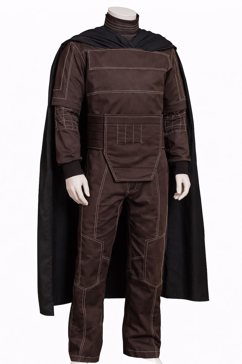 Puede incluir: Un disfraz marr&oacute;n con costuras blancas, con cuello alto, mangas largas y pantalones a juego. Una capa negra est&aacute; drapeada sobre los hombros. El disfraz tiene un dise&ntilde;o estructurado con cintur&oacute;n y detalles acolchados.