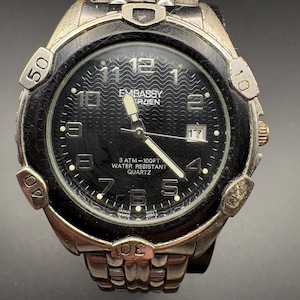 Op de afbeelding: Een zilverkleurig Embassy Ruien quartz horloge met een zwarte wijzerplaat. Het horloge heeft een datumweergave, lichtgevende wijzers en een getextureerde wijzerplaat. De bezel heeft markeringen voor 10, 20, 30, 40 en 50. Het horloge is waterdicht tot 30,48 meter.
