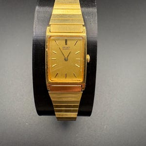 May include: A gold-toned Seiko Quartz watch with a rectangular face and a matching gold band. The watch has a gold dial with black hands and markers. The watch is displayed on a black stand.