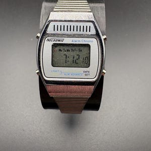 May include: A silver Nelsonic digital watch with a rectangular face showing the time 7:12:10. The watch has a metal strap and features the text "Alarm Chrono" and "LIGHT" and "DATE SET". The watch is displayed on a black stand.