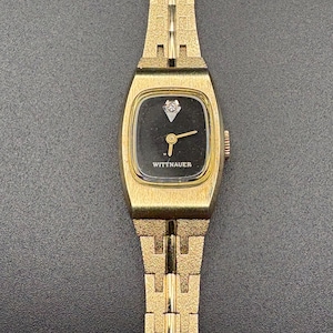 May include: A gold-toned Wittnauer wristwatch with a rectangular black face and a small diamond accent. The watch has a gold band with a textured design. The watch face displays the Wittnauer brand name.