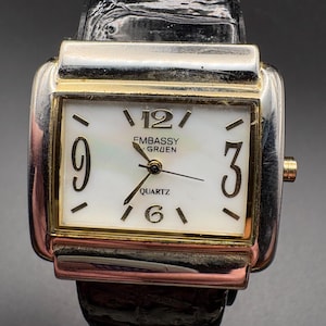 May include: A rectangular Embassy Gruen quartz watch with a white face, gold-tone bezel, and silver-tone case. The watch features black numerals and hands, and a black leather band. The watch face displays the word "QUARTZ".