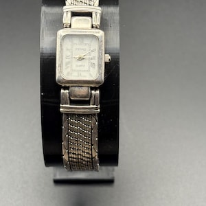 Puede incluir: Un reloj PEDRE de cuarzo plateado con esfera rectangular y números romanos. El reloj presenta una correa de metal tejido y una pequeña corona en el lado derecho. El reloj se exhibe sobre un soporte negro.