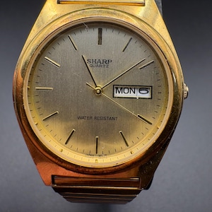 May include: A gold-toned Sharp Quartz watch with a rectangular face. The watch face is a light gold color with black hands and markers. The day and date are displayed. The watch band is also gold-toned.