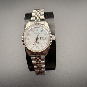 Puede incluir: Reloj Armitron plateado con esfera blanca y correa plateada. La esfera del reloj tiene marcadores con forma de diamante y muestra el día y la fecha. El reloj está sobre un soporte negro.