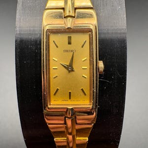 May include: A gold-toned Seiko wristwatch with a rectangular face and a matching gold band. The watch face is gold with black hands and markers. The watch has the word "SEIKO" printed on the face.