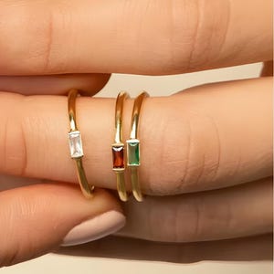 Puede incluir: Tres anillos dorados con piedras preciosas rectangulares claras, rojas y verdes. Los anillos están en un dedo, mostrando el diseño y las variaciones de color de la joyería. Las piedras están engastadas en un estilo minimalista.