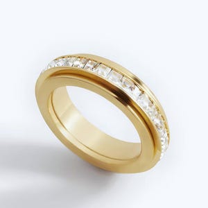 Può includere: Un anello color oro con una fascia di pietre preziose chiare, tagliate a forma quadrata. L'anello ha una finitura liscia e lucida e un design classico. Le pietre sono incastonate in una montatura a canale, creando una linea continua di brillantezza.