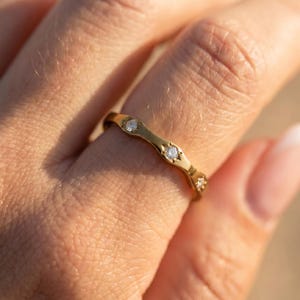 May include: A gold ring with a wavy design, set with small, clear gemstones. The ring is worn on a finger, and the close-up shot highlights the ring's details and the warm tones of the gold.