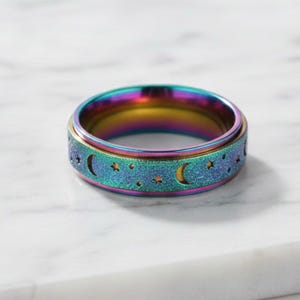 Op de afbeelding: Een veelkleurige spinnerring met een hemels ontwerp. De ring heeft een band met uitgesneden halve manen en sterren. De buitenste band van de ring heeft een getextureerde, glinsterende uitstraling.