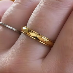 Può includere: Un anello d'oro con un motivo geometrico a diamante. L'anello è di un colore oro brillante e lucido. Nell'immagine è visibile anche un anello d'argento.