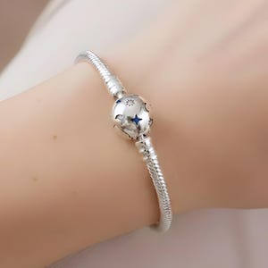 Puede incluir: Una pulsera plateada con un dije redondo con un diseño celestial con detalles de estrellas azules. La pulsera tiene un diseño de cadena de serpiente y se asegura con un cierre. El dije tiene un patrón detallado de estrellas y otros elementos celestiales.