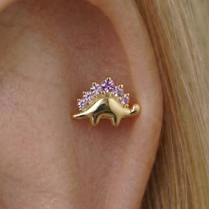 Può includere: Orecchino a forma di dinosauro color oro con una fila di pietre preziose viola sulla schiena. L'orecchino è mostrato su un orecchio, evidenziando il design e il colore del gioiello.